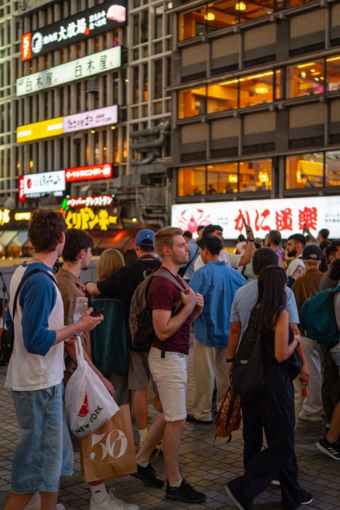 Groepsreis Japan: is een georganiseerde reis iets voor jou? – Kitsune Travel Een menigte mensen staat 's nachts op een stadsstraat met verlichte gevelreclames.