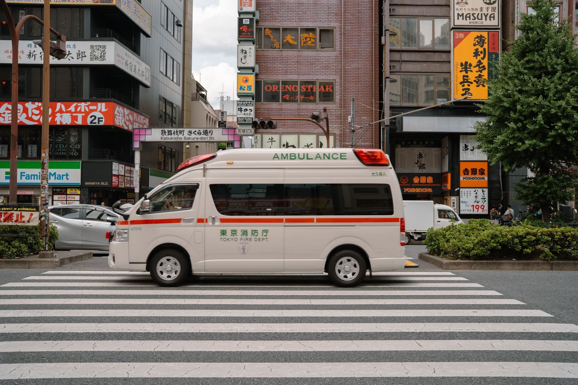 Dokter nodig in Japan? Dit is wat je doet – Kitsune Travel Een drukke straat in Tokyo met een ambulance die een zebrapad oversteekt, omringd door levendige stadsgebouwen.