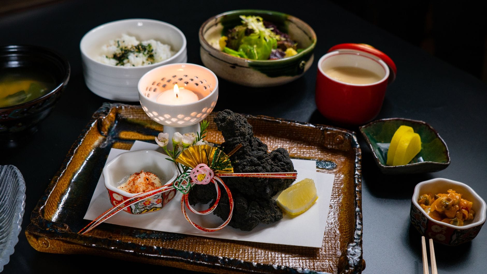 Een artistieke weergave van een Japanse maaltijd met een kaars, diverse gerechten en decoratieve elementen.