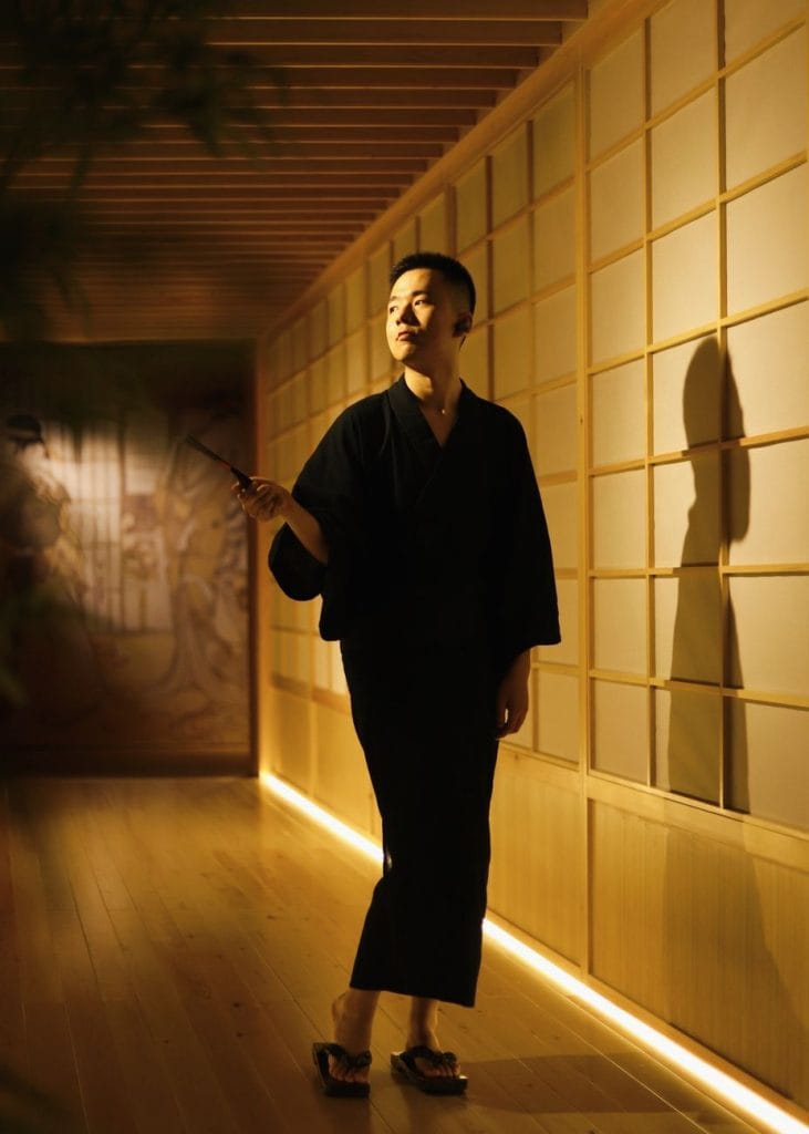 Een Aziatische man in een yukata houdt een voorwerp vast in een warm verlichte, traditionele kamer.