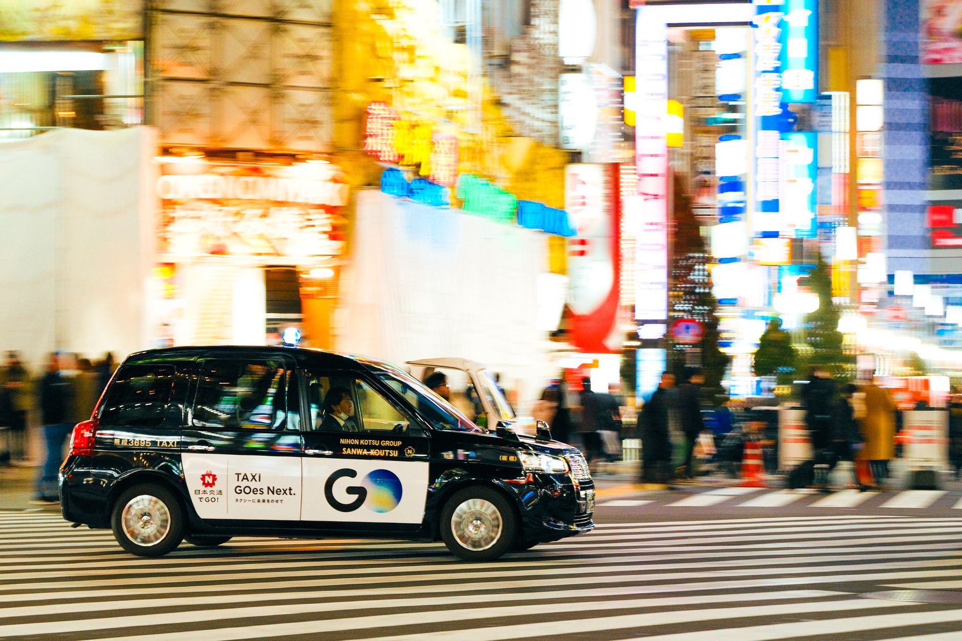 Openbaar vervoer in Tokyo: zo werkt het, van metro tot waterbus – Kitsune Travel Een zwarte taxi rijdt 's nachts door een levendige, kleurrijke stadsstraat onder fel neonlicht.